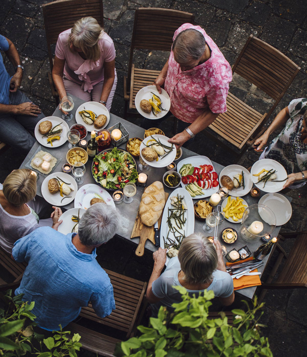 A group of mature friends are sitting around an outdoor dining table, eating and drinking. They are all talking happily and enjoying each others company. The image has been shot from above and taken in Tuscany, Italy.